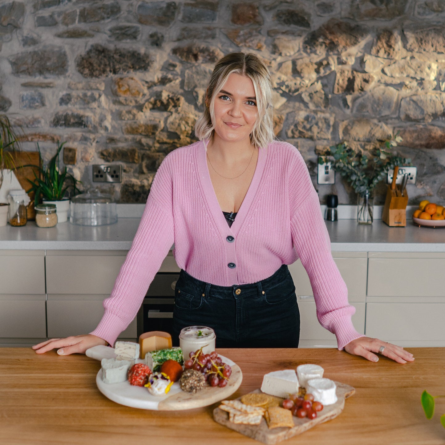 Woman in a pink cardigan standing behind a wooden table with a cheeseboard and platter in a kitchen.