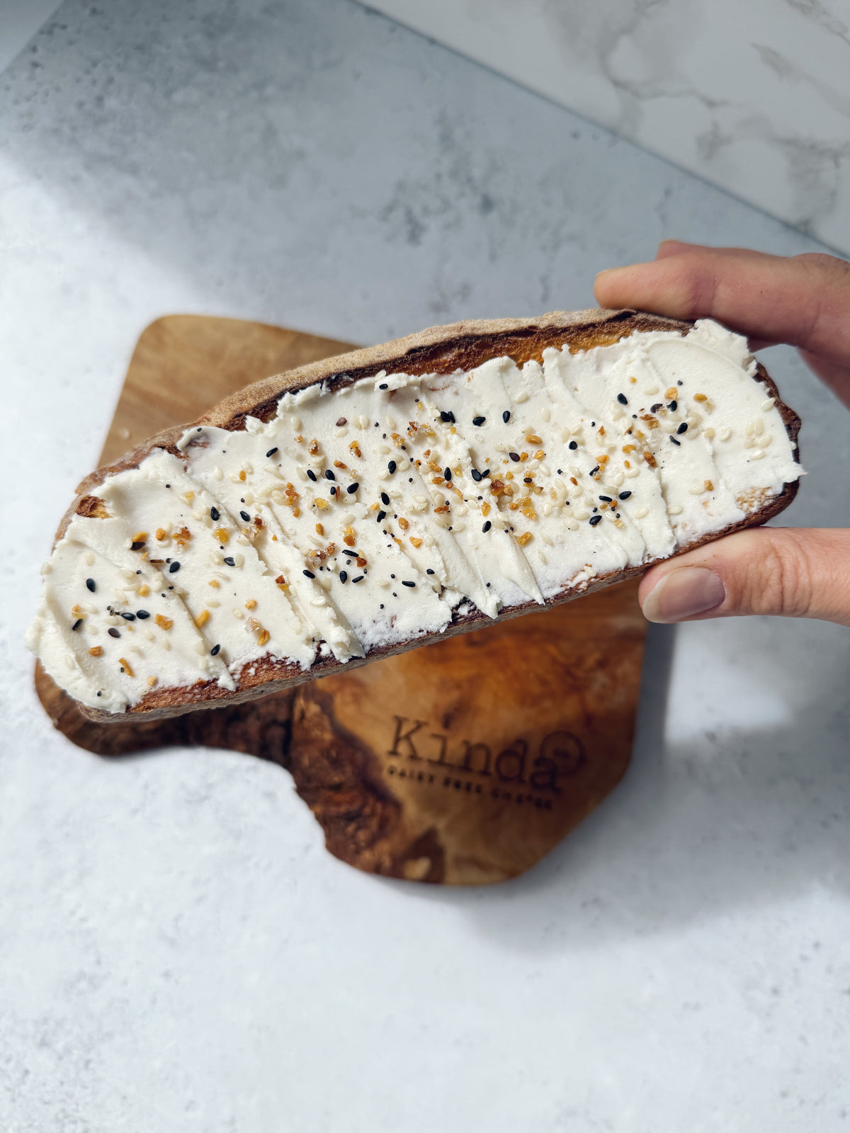 A hand holds a toasted sourdough bread slice spread with thick white cream and sprinkled black and brown seeds, resting over a wooden cutting board branded Kinda Baked Goods.