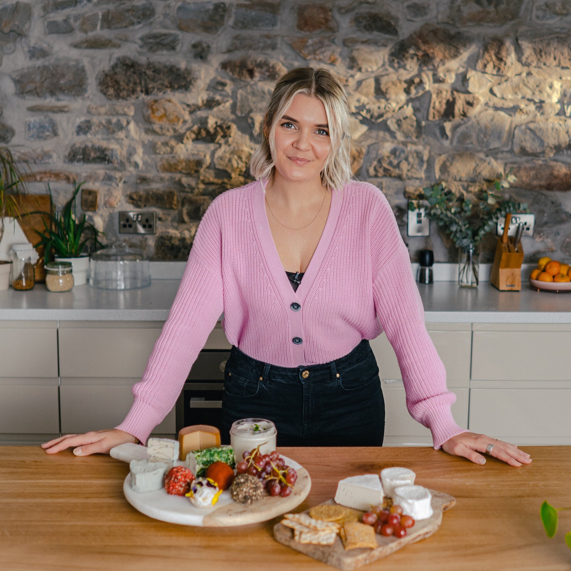 Woman in a pink cardigan standing behind a wooden table with a cheeseboard and platter in a kitchen.